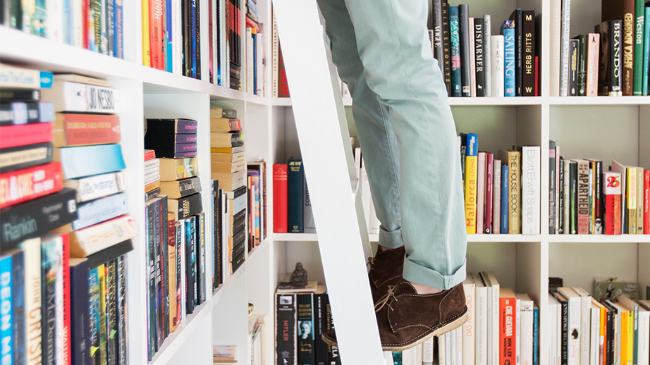 Enkele boekenrekken staan in een hoek tegen elkaar. Tegen een rek staat een ladder. Op die ladder zijn iemand voeten en benen te zien. Het kan in een bibliotheek zijn.