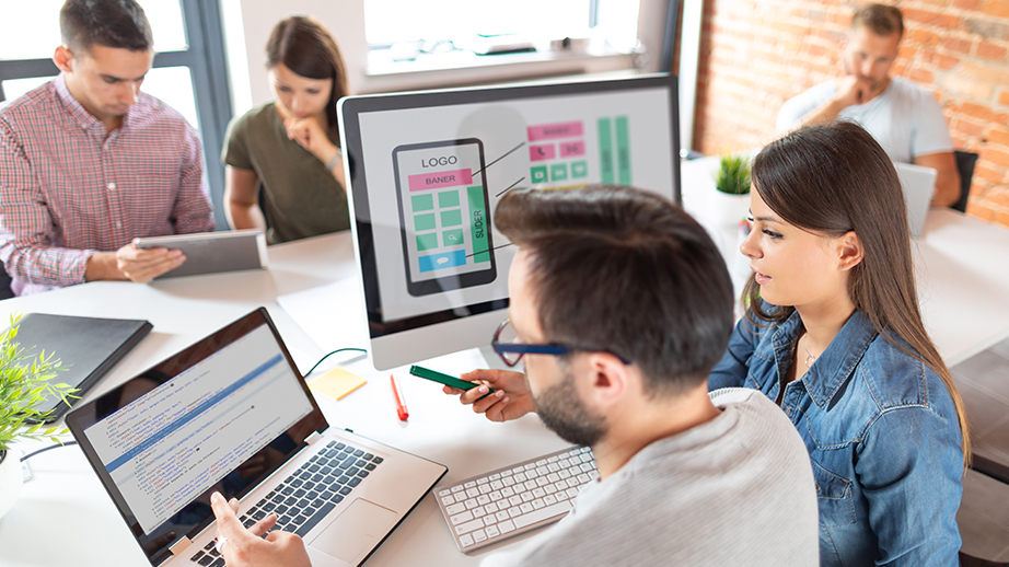 Vijf mensen zitten aan een tafel. Ze werken op computers. Een man en een vrouw zijn iets aan het programmeren. Ze ontwikkelen een app en gebruiken daarvoor software.