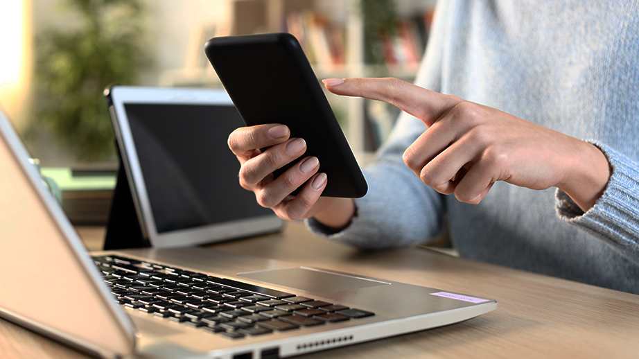 Iemand heeft een smartphone in de rechterhand en wijst er met de linkerwijsvinger naar. Er staan nog een computer en een tablet op tafel.