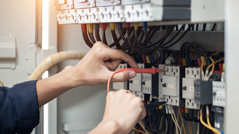 Twee handen hebben de rode en de zwarte kabel van een multimeter vast. Daarmee checkt iemand of de aansluitingen in de elektriciteitskast in orde zijn. In die elektriciteitskast zijn veel kabels en zekeringen.