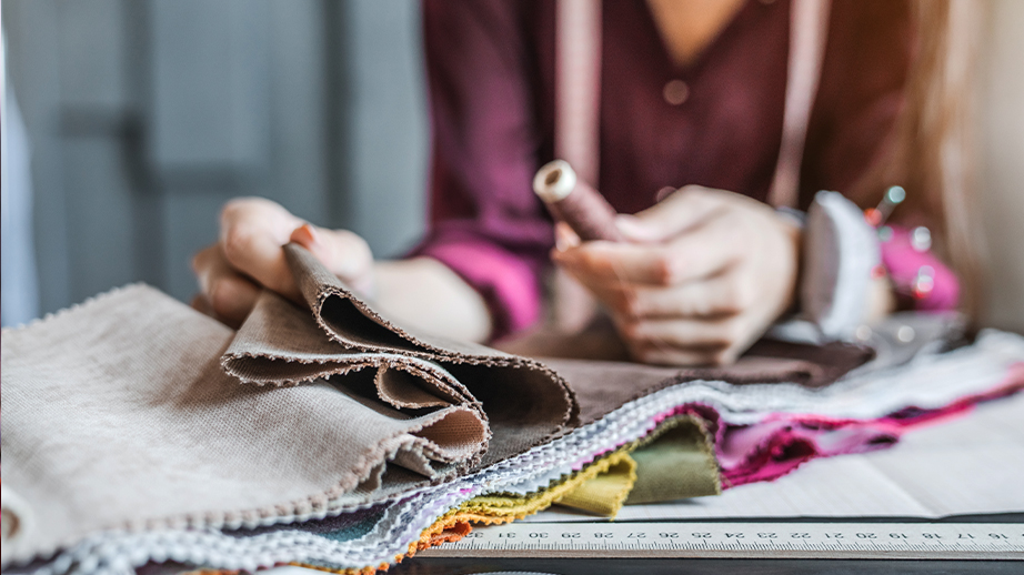 Op een tafel liggen effen stoffen in allerlei kleuren: beige, bruin, wit, geel, groen en roos. Onder de stoffen ligt patroonpapier en een lintmeter. Iemand heeft een bobijn stikgaren vast en heeft op de linkerpols een speldenkussen. Dit beeld doet denken aan mode, interieur en ontwerp.