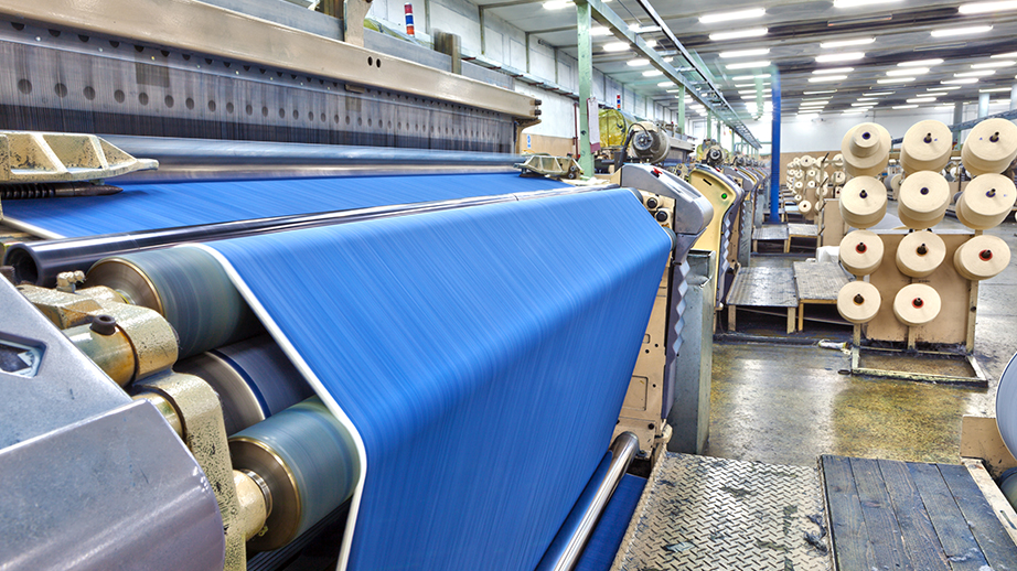 Dit is een fabriek voor textielproductie. Er worden stoffen gemaakt. Op de voorgrond staat een grote machine waar een blauwe stof tui rolt. Erachter staan nog enkele andere machines. Bij elke machine staan bobijnen met touw of wol.