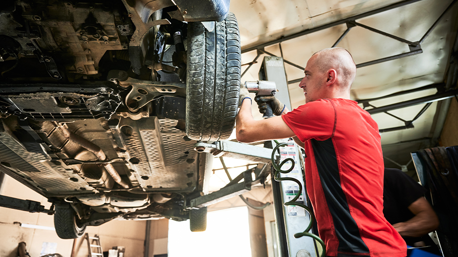 Een kale man in een rood T-shirt werkt aan een auto. Die auto is opgehangen en de onderkant is zichtbaar. De wielen van de wagen zijn op ooghoogte van de man. De man maakt met een schroeftoestel de bouten van de wielen vast. Alles speelt zich af in een werkplaats voor voertuigen en fietsen.