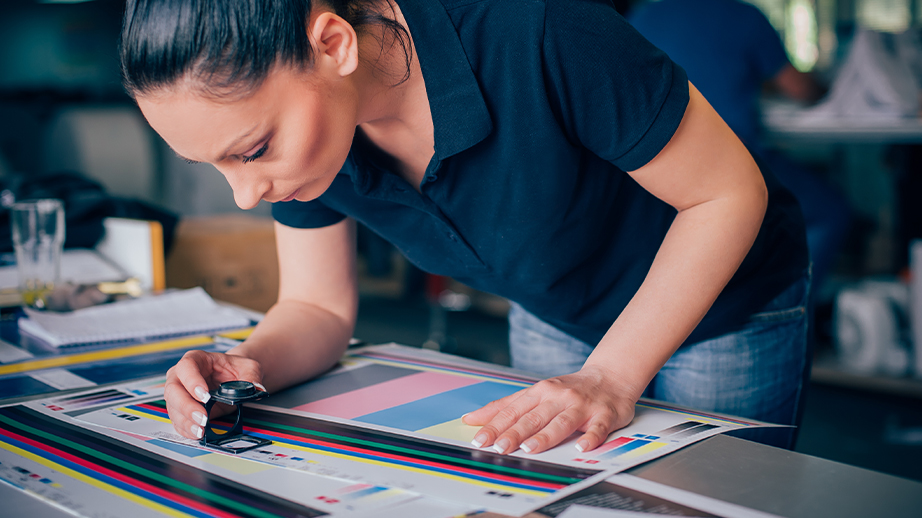 Een vrouw staat voorovergebogen aan een tafel. Ze bekijkt met een vergrootglas een drukproef. Het is in een werkplaats voor grafische communicatie en drukwerk.